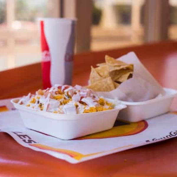 A white bowl of loaded nachos with a side of tortilla chips and a soda.