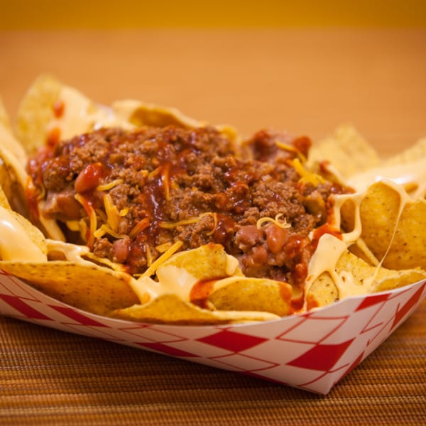 Chili cheese nachos in a red and white checkered paper tray, on a brown woven mat.
