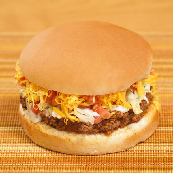 A close-up of a taco burger with ground beef, shredded cheese, lettuce, tomatoes, and sour cream.